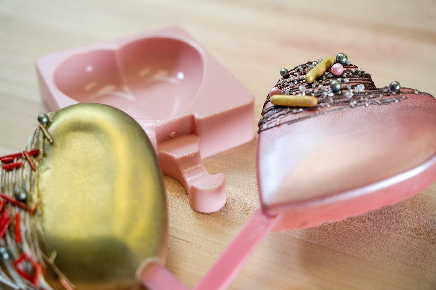 Heart-shaped chocolate pops with sprinkles and a pink silicone mold on a wooden surface.