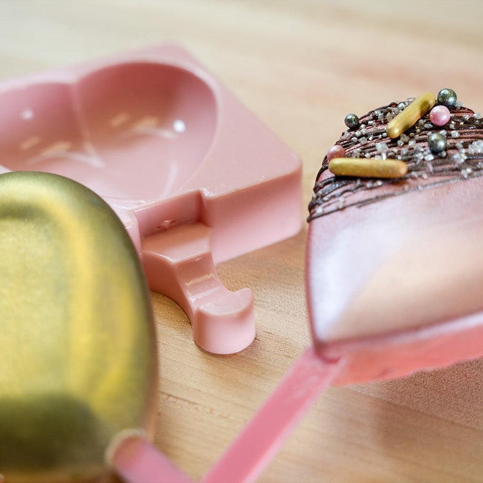 Heart-shaped chocolate pops with sprinkles and a pink silicone mold on a wooden surface.