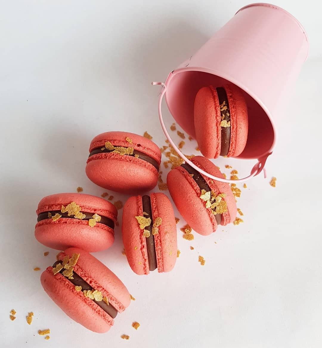 Pink macarons with chocolate filling and gold flakes spilling from a decorative pink bucket.