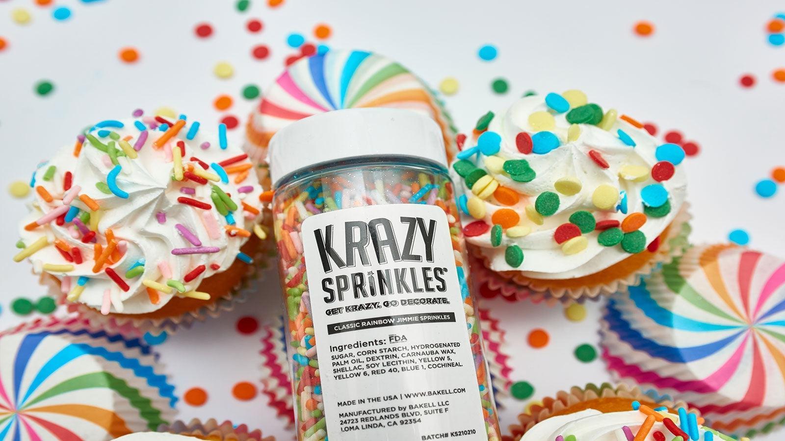 A clear bottle of Krazy Sprinkles, classic rainbow jimmie sprinkles, surrounded by colorful cupcakes.