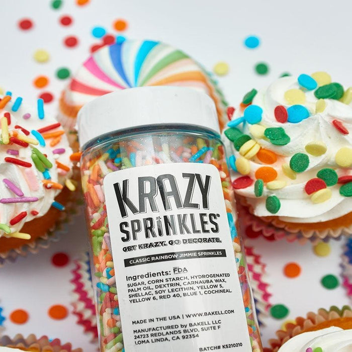 A clear bottle of Krazy Sprinkles, classic rainbow jimmie sprinkles, surrounded by colorful cupcakes.