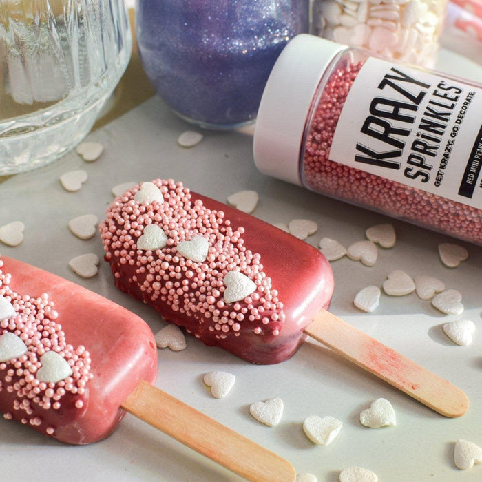 Two red popsicles decorated with pink sprinkles and white heart candies, next to a Krazy Sprinkles container.