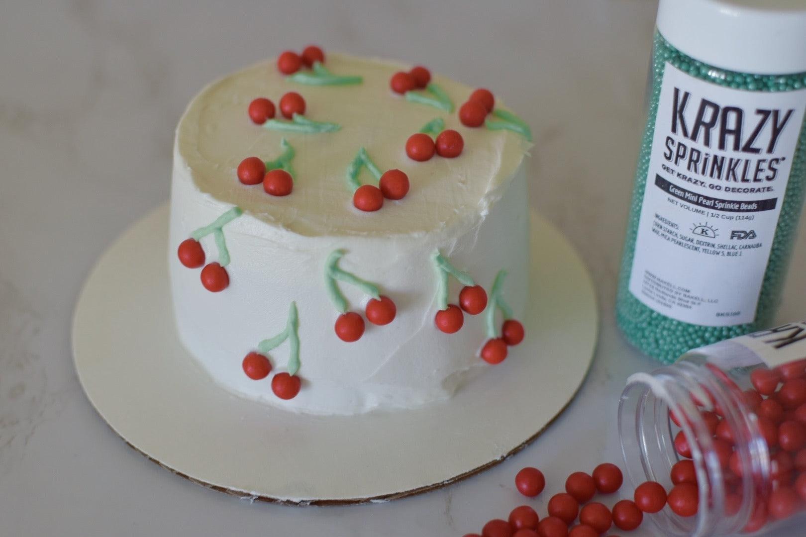 White frosted cake decorated with red cherry candies and green icing stems, next to a bottle of green sprinkles.