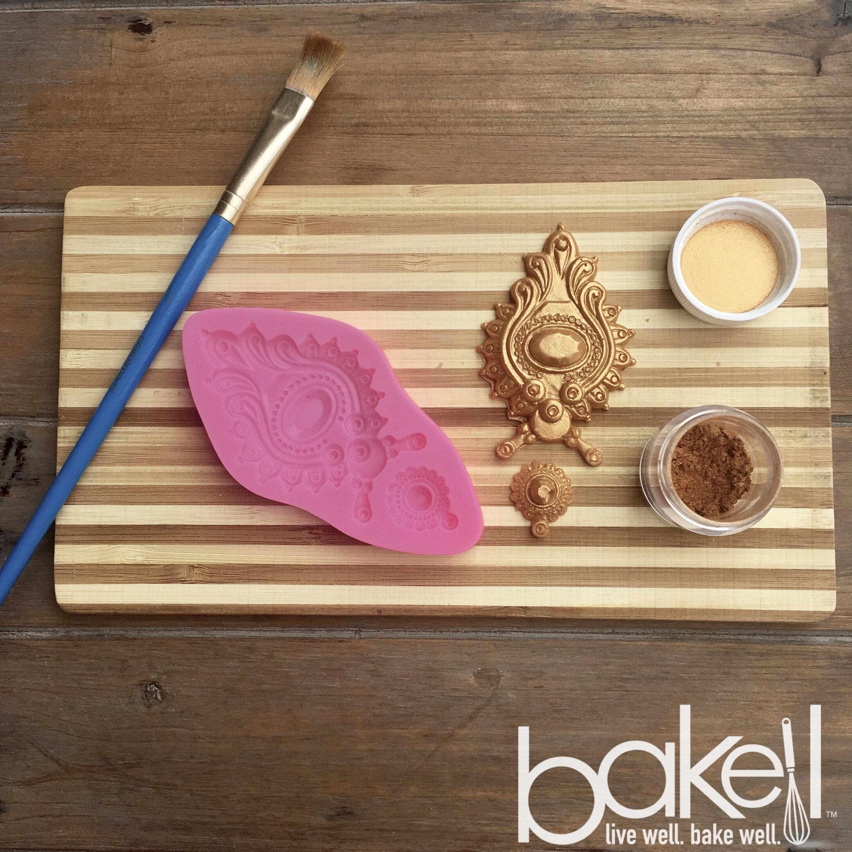 A cutting board, with a paintbrush, design tools, and bronze Luster Dust on top of the cutting board. | bakell.com