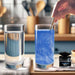 Hand stirring blue edible Brew Glitter into a glass of water next to a 4g jar on a kitchen counter.