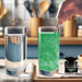 Hand stirring green color-changing brew glitter into a clear drink, next to a plain glass of water and a glitter jar.