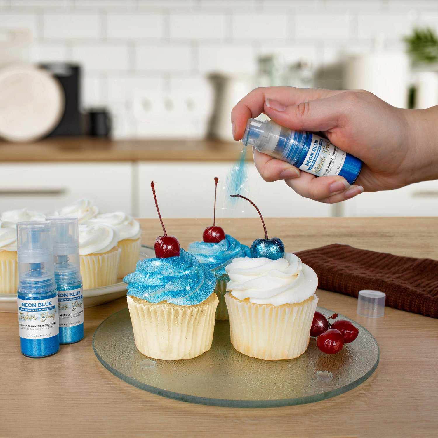 Hand spraying Neon Blue Tinker Dust edible glitter from a 4g pump onto cupcakes with cherries.