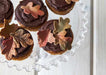 Chocolate cupcakes topped with edible autumn leaves in olive green, brown, and bronze, on a clear scalloped cake stand.