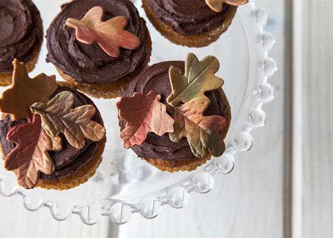 Chocolate cupcakes topped with edible autumn leaves in olive green, brown, and bronze, on a clear scalloped cake stand.