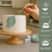 Hand painting an olive green leaf design on a white cake with luster dust, alongside product container and "Mix, Paint, Shine" icons.