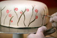 Gloved hands decorating a white cake with delicate brown branches and pink flowers using a fine brush.
