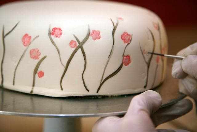 Gloved hands decorating a white cake with delicate brown branches and pink flowers using a fine brush.