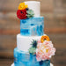 Four-tier wedding cake with Patriot Blue Luster Dust, white tiers, and colorful floral decorations.