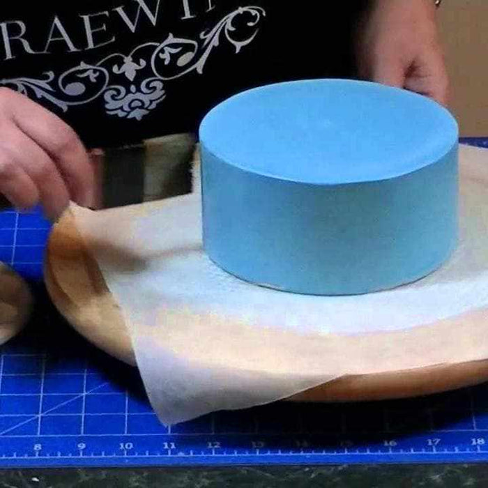 Hands applying periwinkle blue luster dust to a round cake on parchment paper.