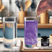 Hand stirring purple color-changing brew glitter into a clear drink, next to a plain glass of water and a small jar of glitter.