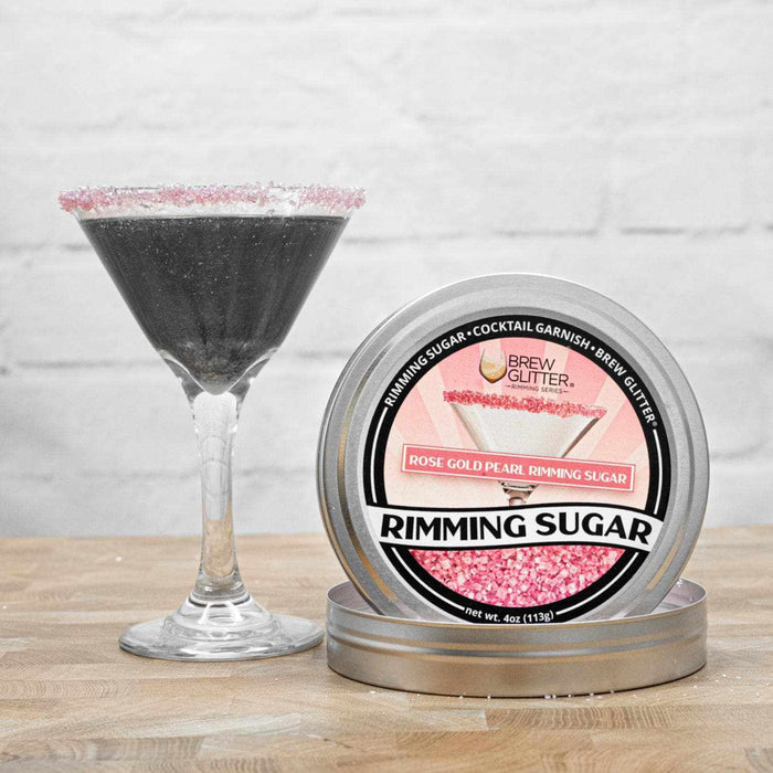 Rose gold pearl rimming sugar tin next to a martini glass with a black drink and pink sugar rim.