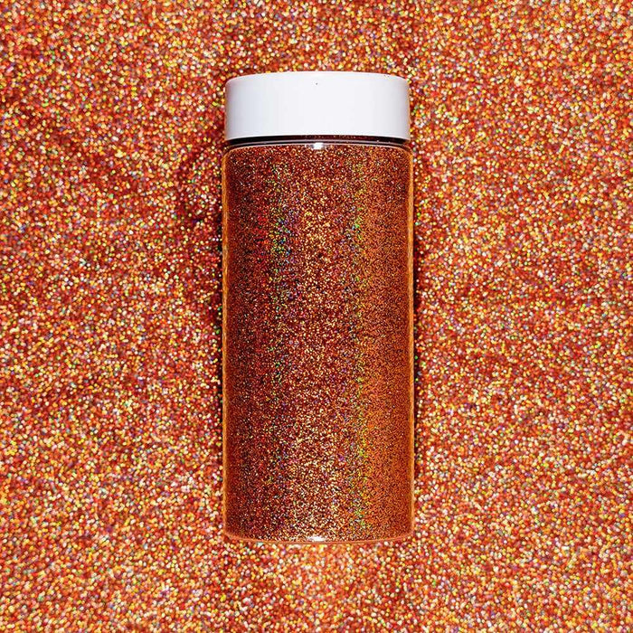 Rose gold ultrafine holographic craft glitter in a clear jar with a white lid, on a matching glitter background.