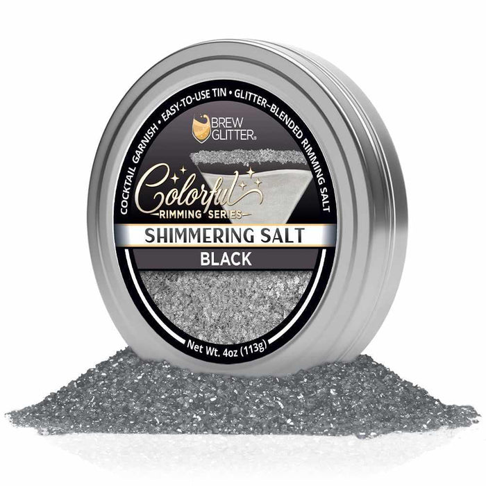 Silver tin of shimmering black cocktail rimming salt with a labeled lid, resting in a pile of coarse black salt crystals.
