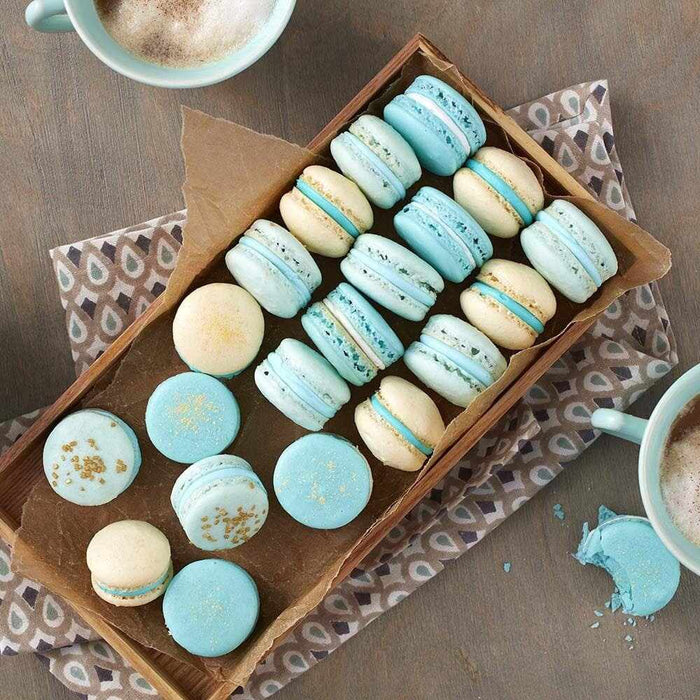 Soft blue and cream macarons with gold luster dust, arranged in a wooden tray with coffee cups.