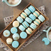 Soft blue and cream macarons with gold luster dust, arranged in a wooden tray with coffee cups.