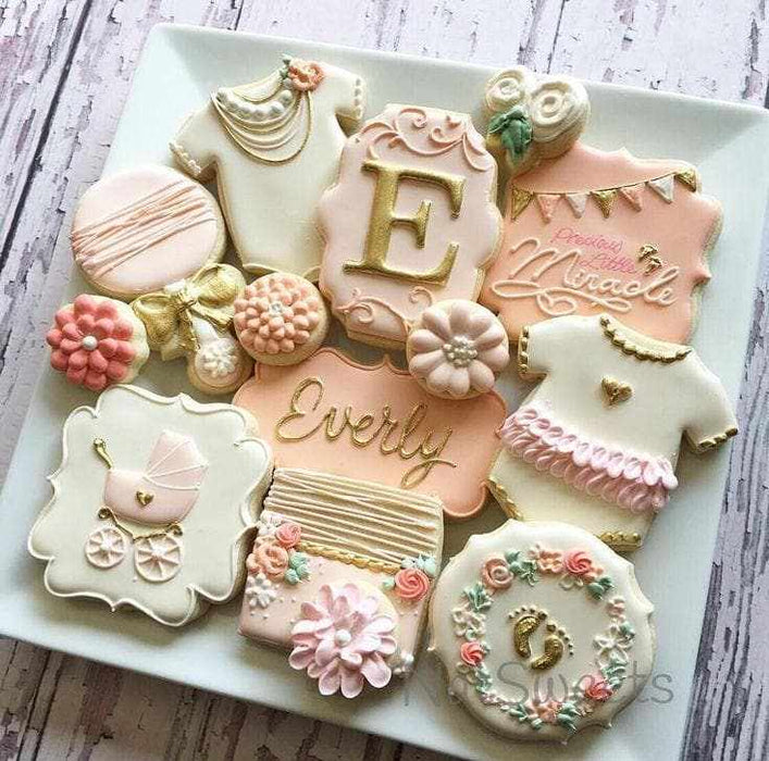 Assortment of decorated baby shower cookies in pink, white, and gold, featuring onesies, a stroller, and "Everly".