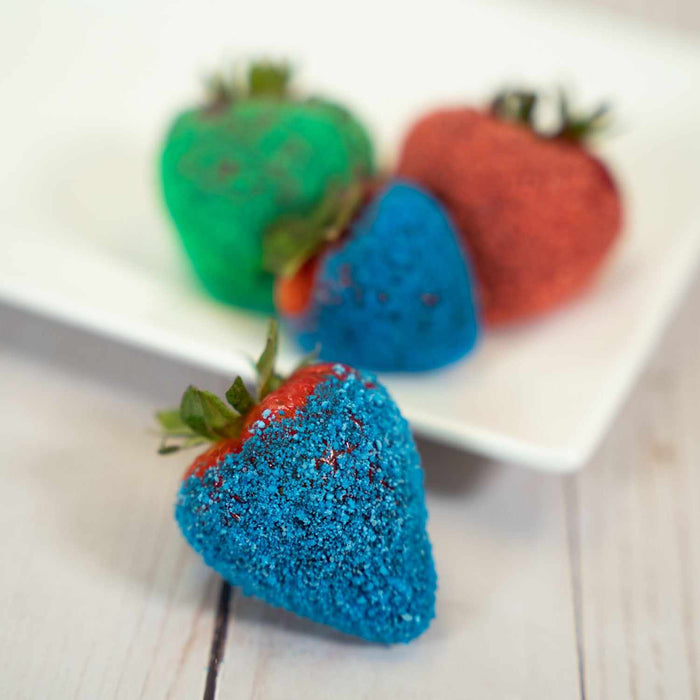Fresh strawberries coated in vibrant blue, green, and red Sour Blue Raspberry candy toppers on a white plate.