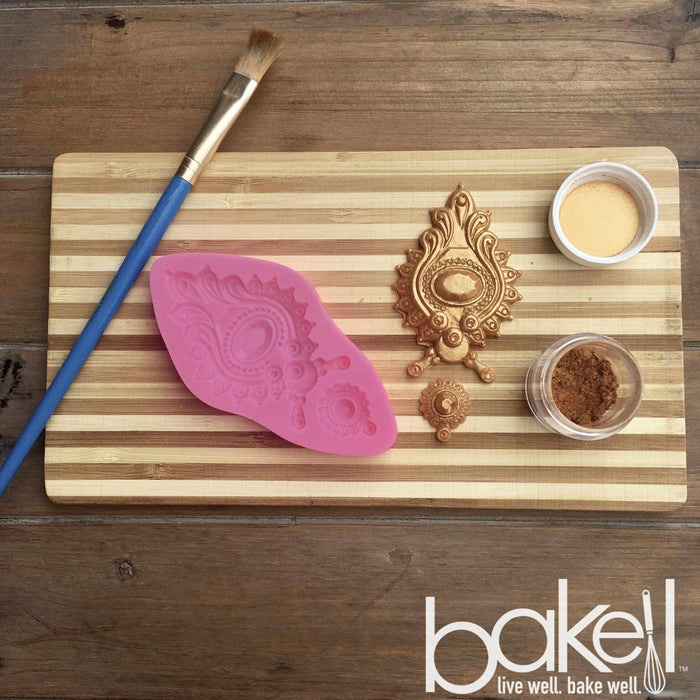 A cutting board, with a paintbrush, design tools, and bronze Luster Dust on top of the cutting board. | bakell.com