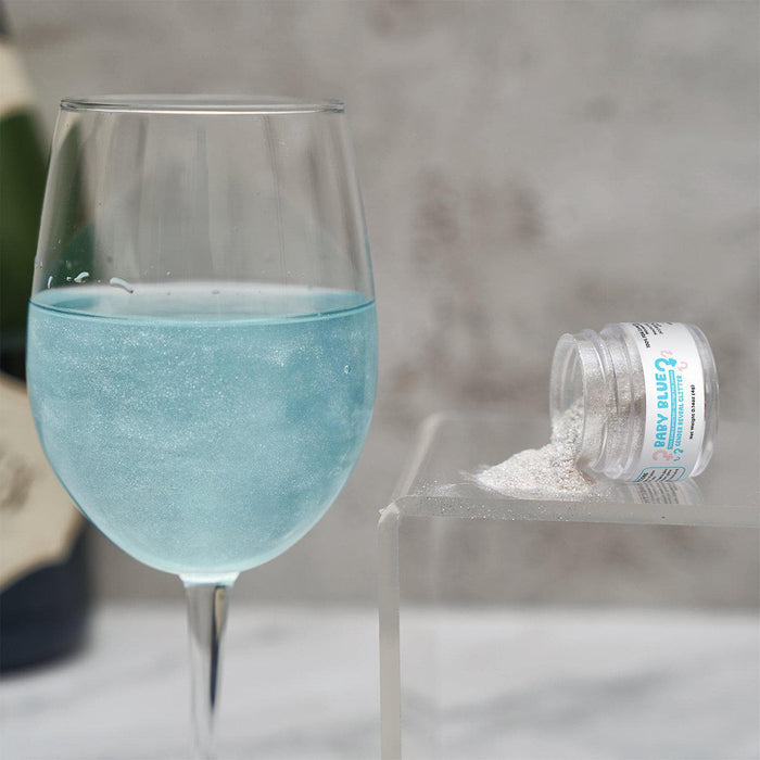 Wine glass filled with shimmering baby blue drink next to a small jar of white edible gender reveal glitter.
