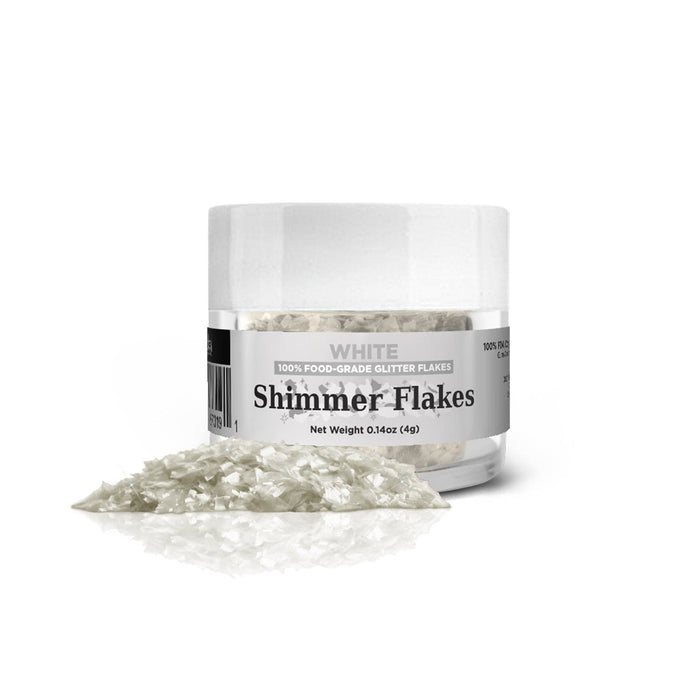 White edible shimmer flakes in a clear 4g jar with some flakes spilled, reflecting on a white surface.