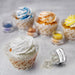 White edible shimmer flakes spilling from a jar next to a frosted cupcake, with other colorful cupcakes and flakes in the background.