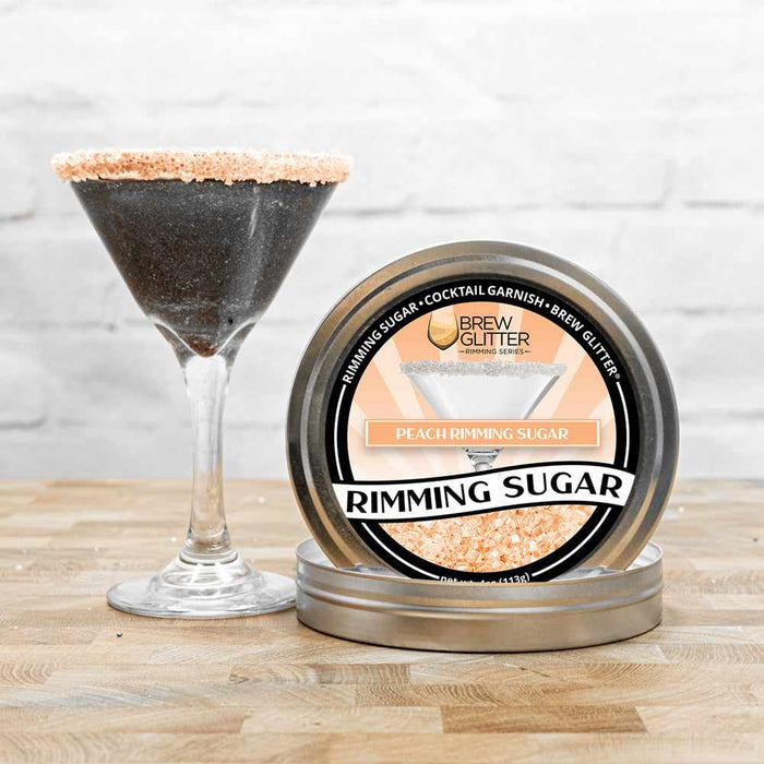 A front view of two items:  To the left, a glass filled with black  and glittery liquid, and Peach  Rimming Sugar on top.  To the right, a can of Peach Rimming Sugar facing forward, and placed in the bottom of a rim jar. | bakell.com
