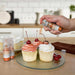 Three cupcakes being sprayed by a Pumpkin Orange color Edible Glitter 4 gram pump. | bakell.com