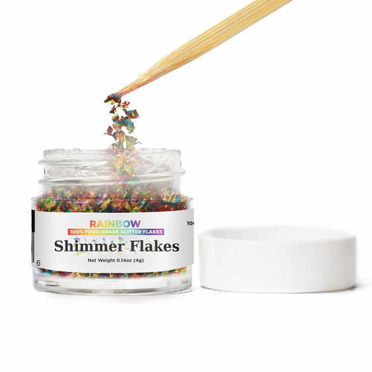 Front view of an open jar of Rainbow Edible Shimmer Flakes, with a wooden stick stirring the flakes | bakell.com