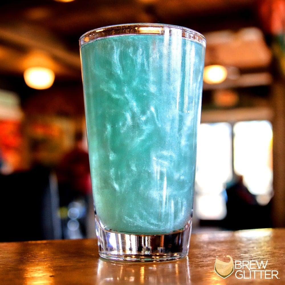 Tall glass with shimmering teal edible glitter in a beverage, on a wooden bar.