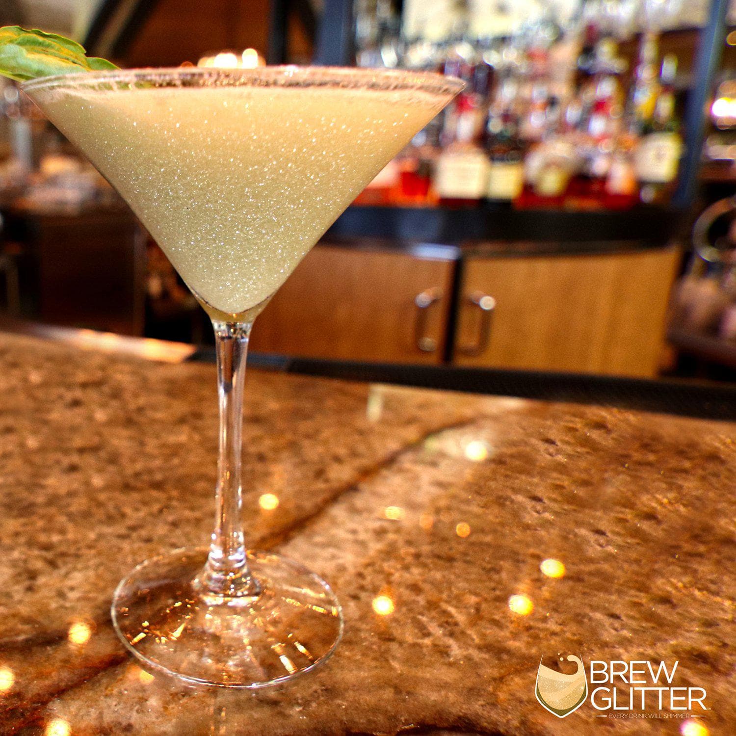 Shimmering light yellow cocktail in a martini glass with a green garnish on a bar counter.