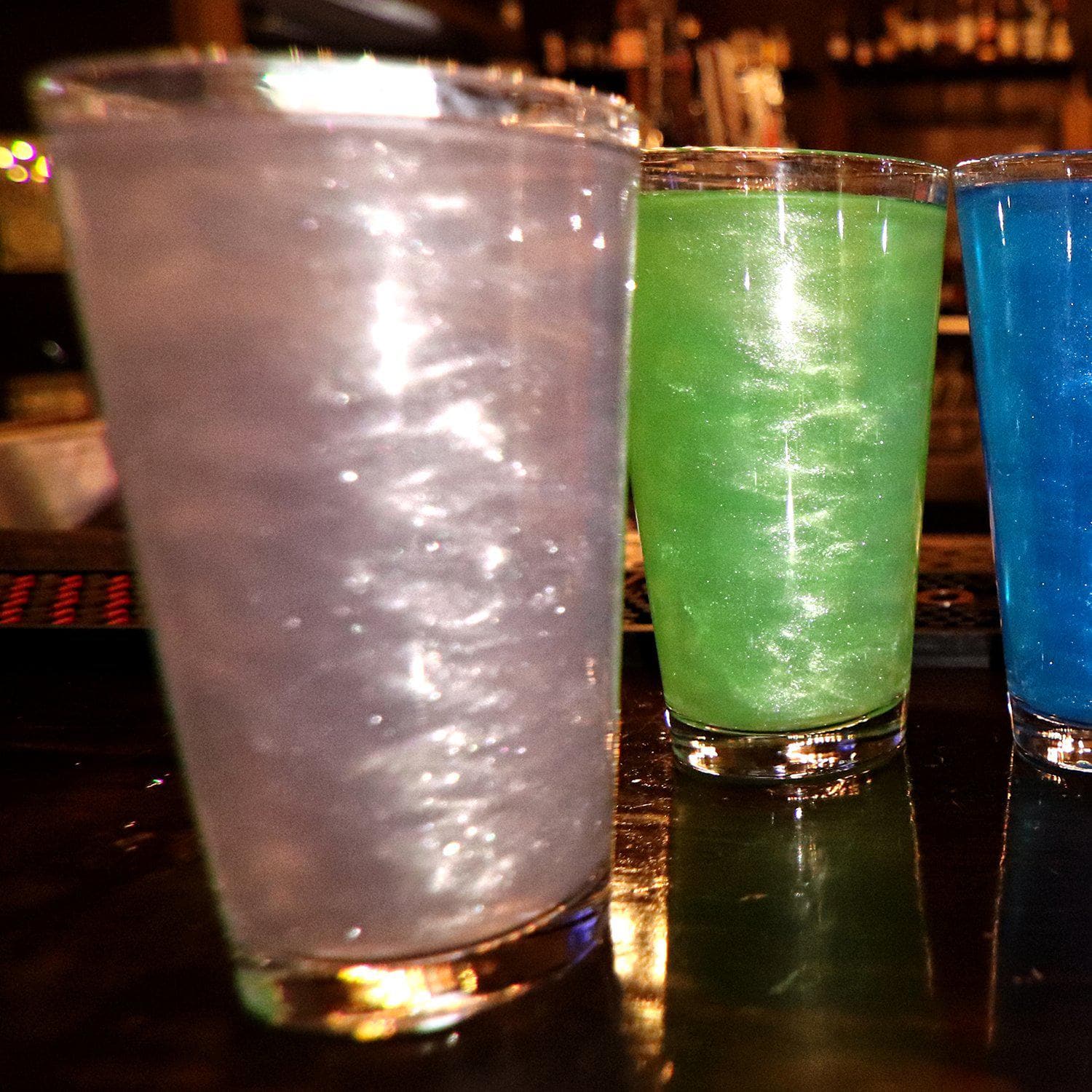 Three tall glasses of shimmering, colorful drinks: purple, green, and blue, on a dark bar.