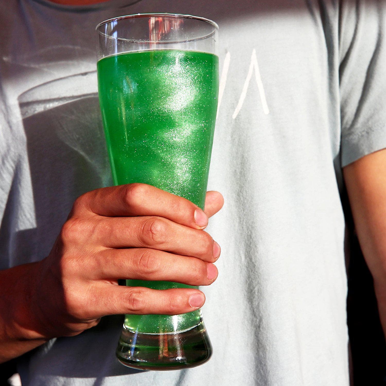 Hand holding a tall glass of shimmering green Brew Glitter drink, perfect for summer beverages.